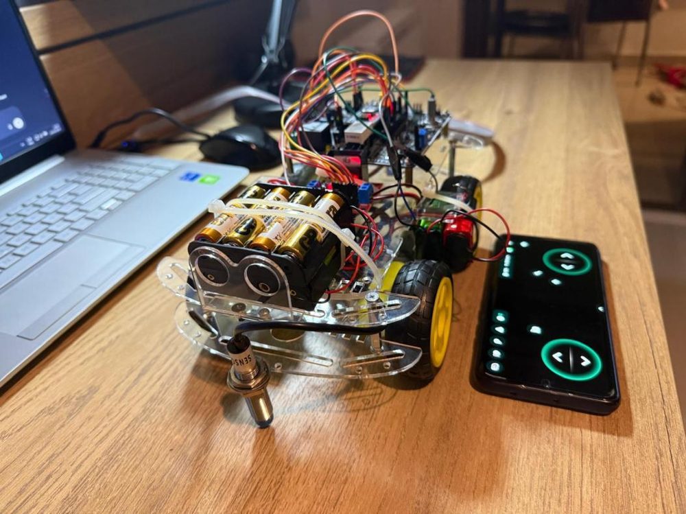 Keşif robotu: şeffaf akrilik şasi, önde pil paketi ve aşağı bakan endüktif sensör, yanında kontrol uygulaması açık telefon